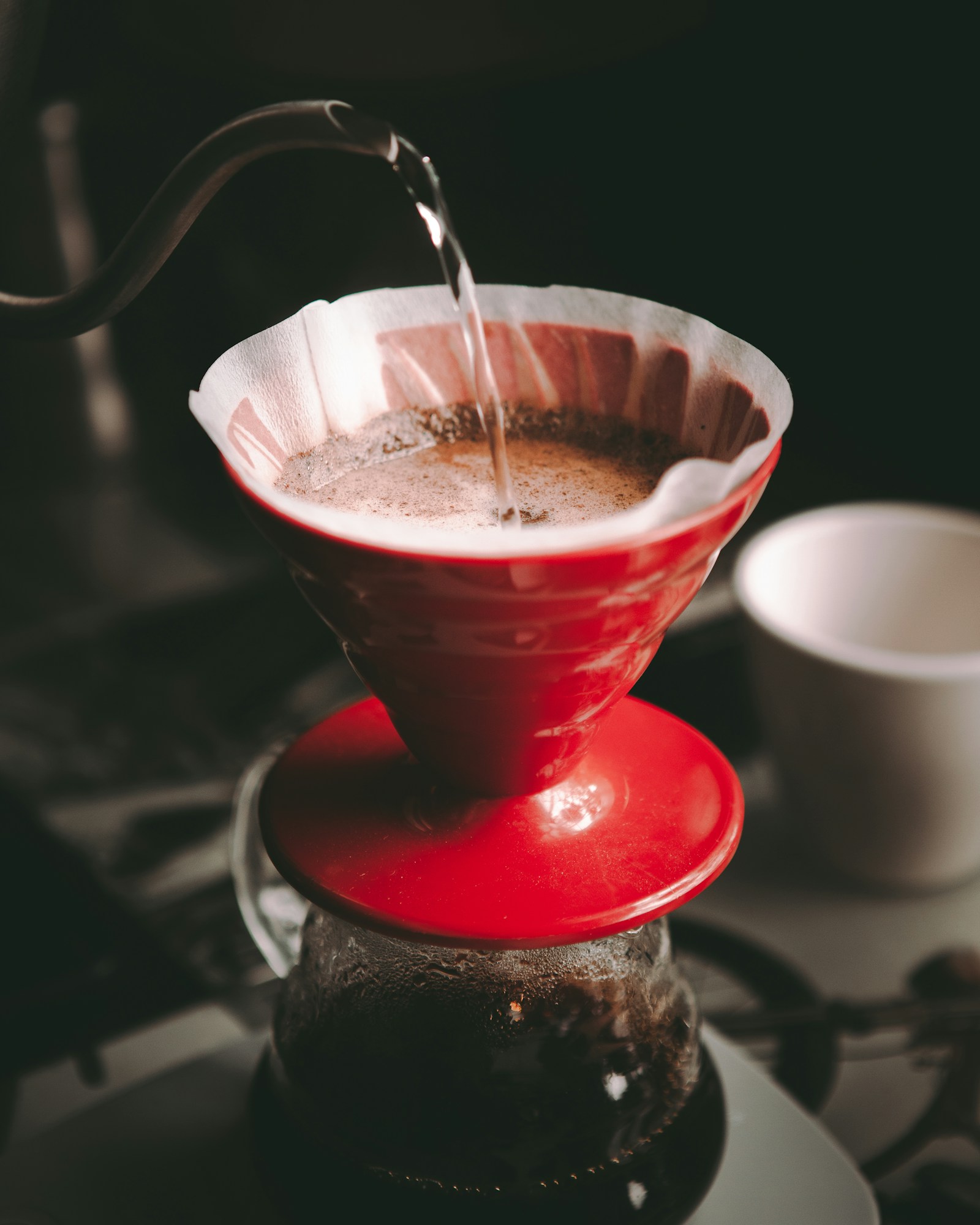 Hot water poured from a gooseneck kettle into a red V60-style dripper with a paper filter